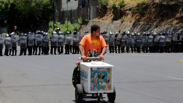 An ice-cream salesman walks in front of riot police agents blocking the road as demonstrators protest during a march to the National Congress to demand justice for the 43 deaths in recent protests in Managua on May 2nd, 2018.
