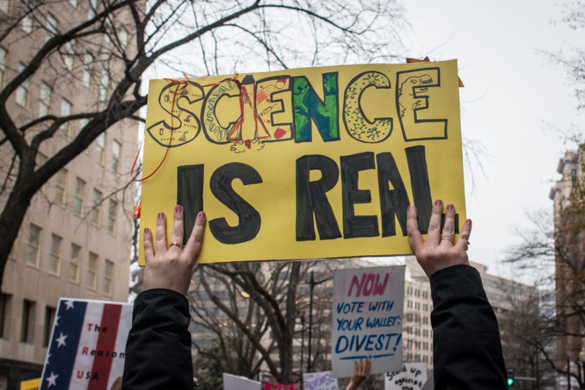 Photo showing hands holding up a sign that reads, "SCIENCE IS REAL"
