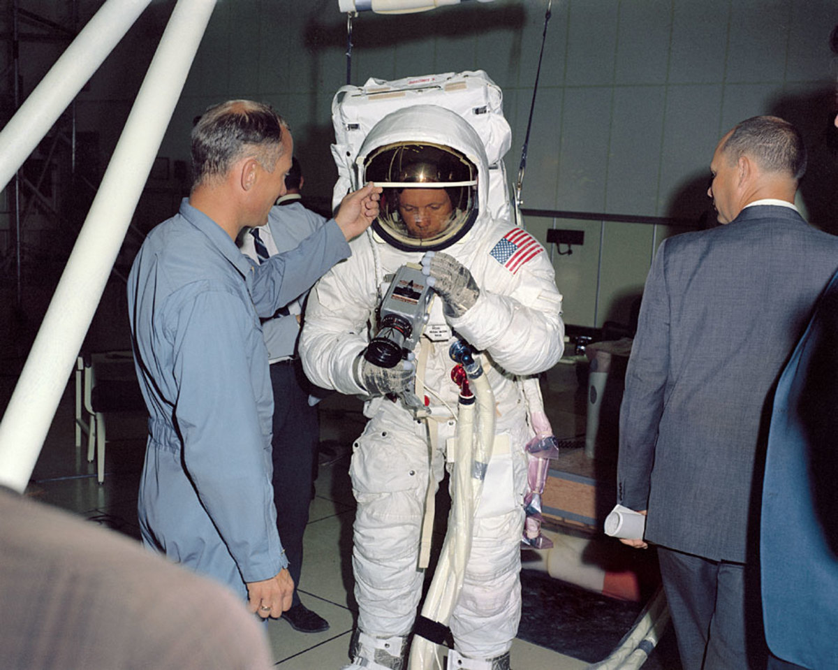 During training, Neil Armstrong practices attaching a Hasselblad camera to a mount on his suit. Buzz Aldrin helps him see what he's doing by raising his visor.