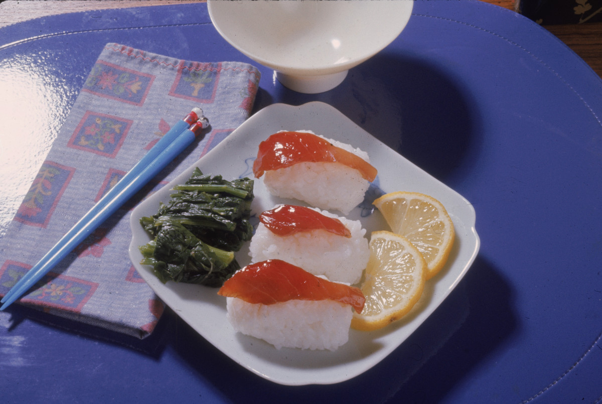 Photograph shows a plate with sushi on rice, slices of lemon, and some green food item, probably seaweed, on a mat with a bowl, napkin, and pair of chopsticks, 1970s.