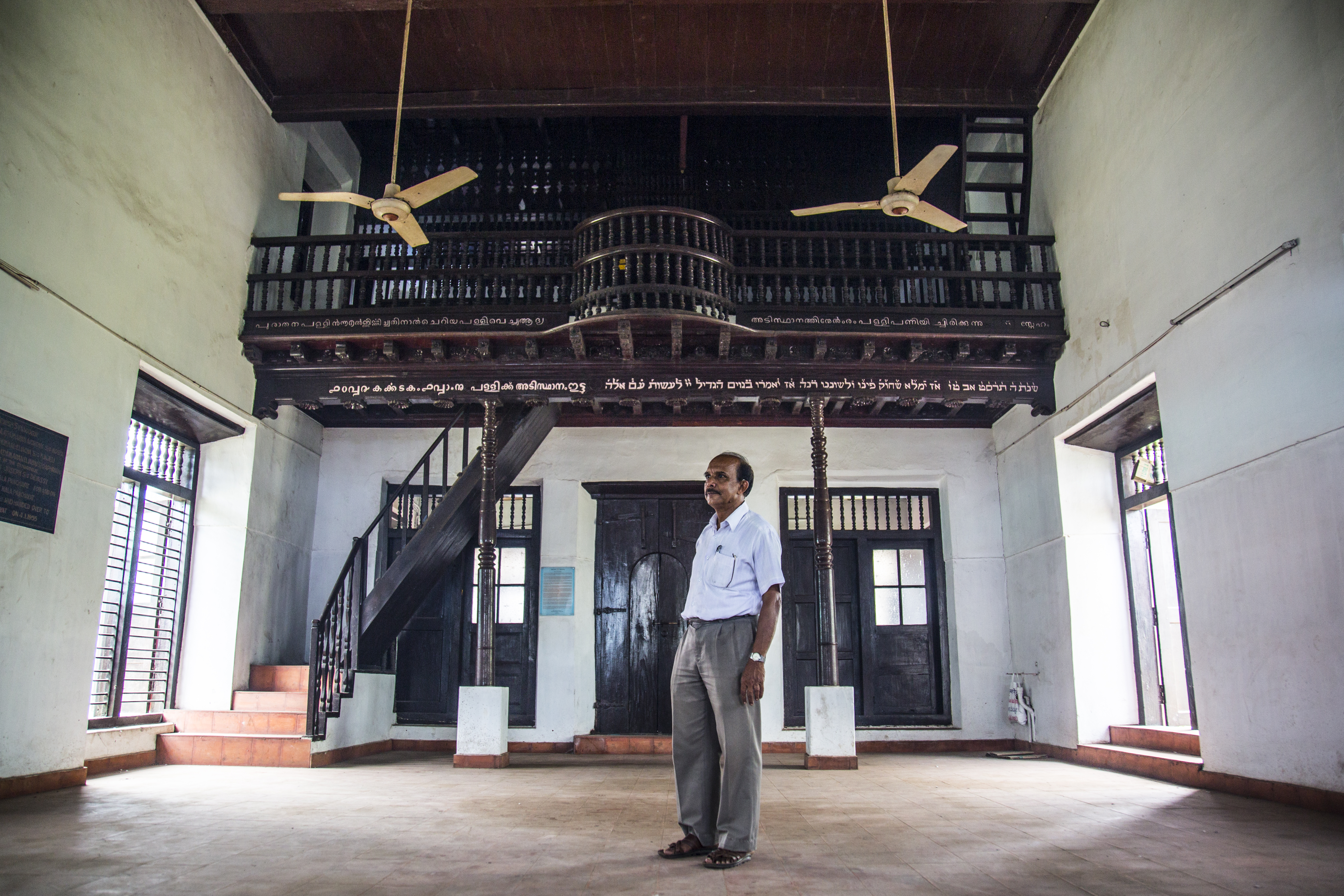 Karma Chandran stands in the empty Mala Synagogue, 50 kilometers from Cochin. He is fighting to preserve the Mala Synagogue and Jewish cemetery, two sites that have been heavily neglected for decades. When the Mala Jewish community migrated to Israel in 1955, they made a written agreement with the local government to protect the sites, but, in 2012, the local government made plans to build a stadium on cemetery land anyway. Now, the blue-and-red stadium looms over the remaining tombstones, the stadium left empty and half-constructed because of ongoing court cases between the government and the departed Jewish community.