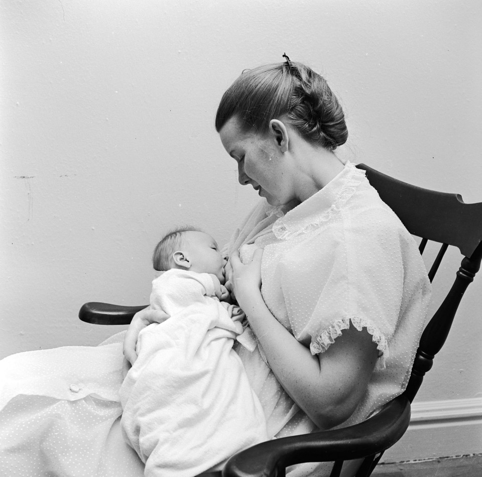 Black-and-white photo showing a woman in a rocking chair, breastfeeding an infant