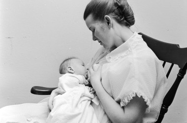 Black-and-white photo showing a woman in a rocking chair, breastfeeding an infant