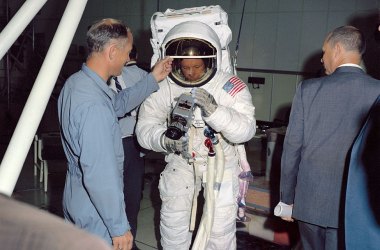 During training, Neil Armstrong practices attaching a Hasselblad camera to a mount on his suit. Buzz Aldrin helps him see what he's doing by raising his visor.