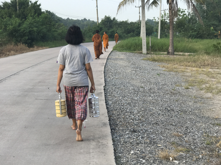 A young Thai girl follows three novice monks in the collection of the morning alms, in which they accept donations of food and drink to the temple from residents throughout the village of Chonburi.