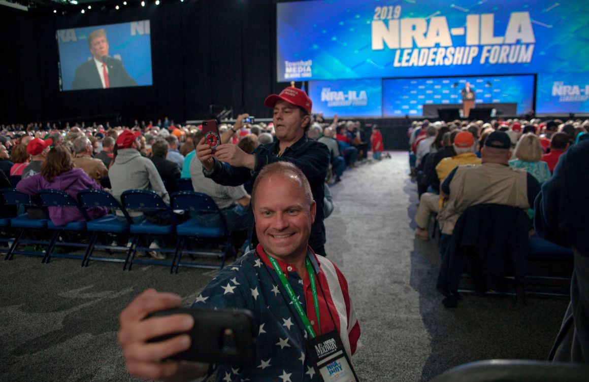 Skippy Thomas, a member of the National Rifle Association, takes a selfie with President Donald Trump in the background during the NRA's annual meeting at Lucas Oil Stadium in Indianapolis, Indiana, on April 26th, 2019.