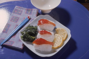 Photograph shows a plate with sushi on rice, slices of lemon, and some green food item, probably seaweed, on a mat with a bowl, napkin, and pair of chopsticks, 1970s.