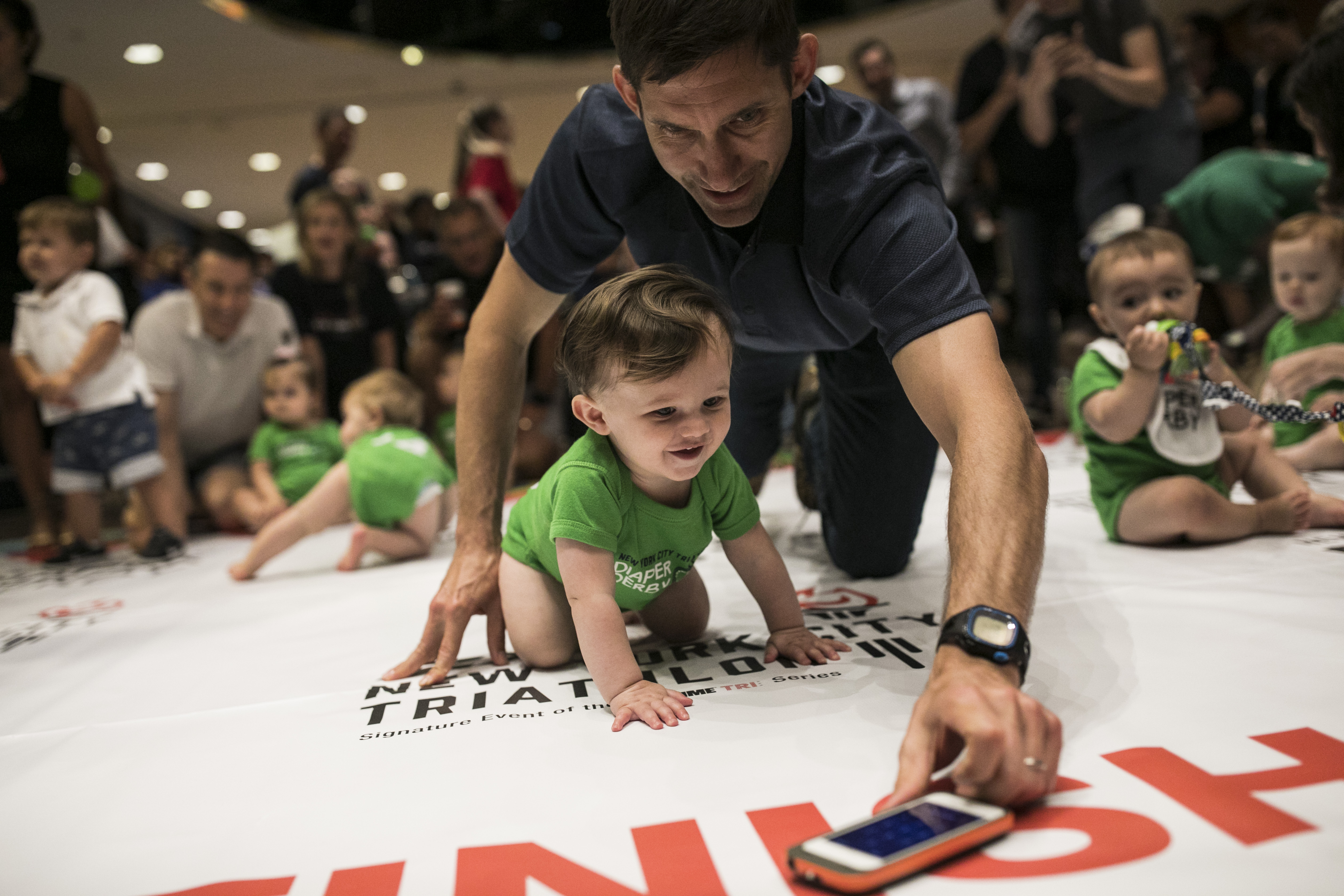 A father lures his child along with an iPhone during warm-ups before babies race in the NYC Triathlon's annual Diaper Derby on July 14th, 2017, in New York City.