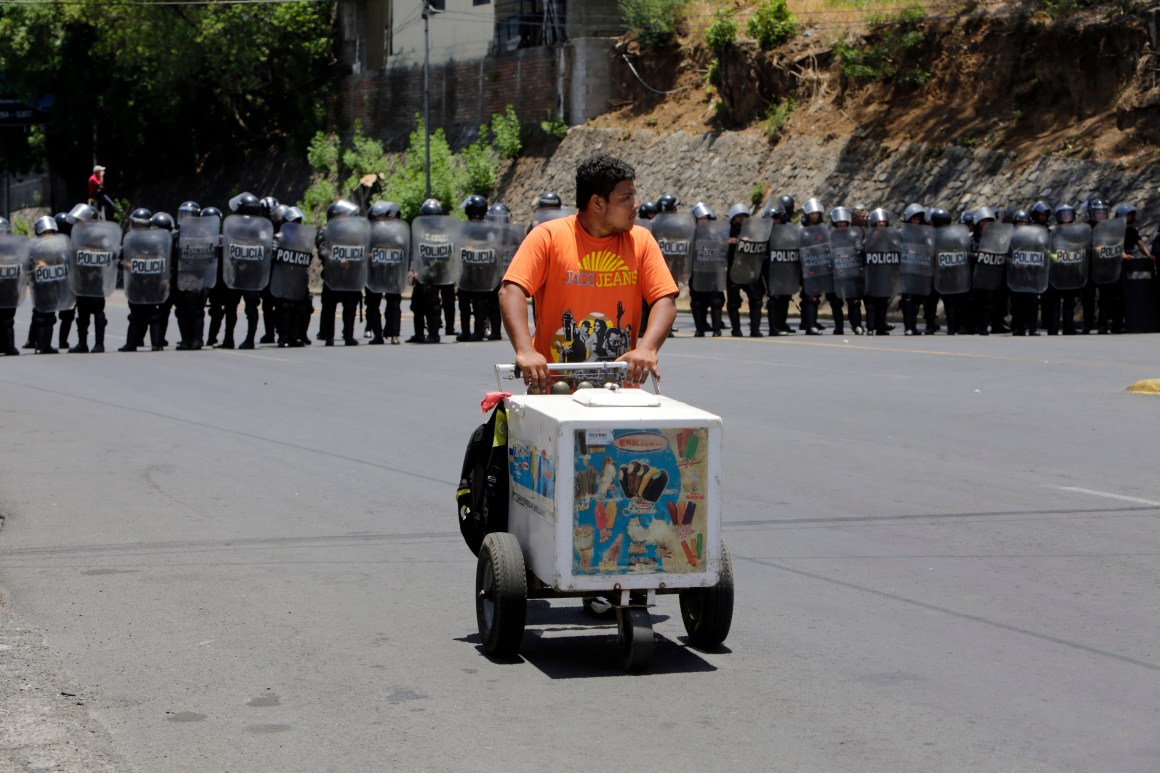 An ice-cream salesman walks in front of riot police agents blocking the road as demonstrators protest during a march to the National Congress to demand justice for the 43 deaths in recent protests in Managua on May 2nd, 2018.