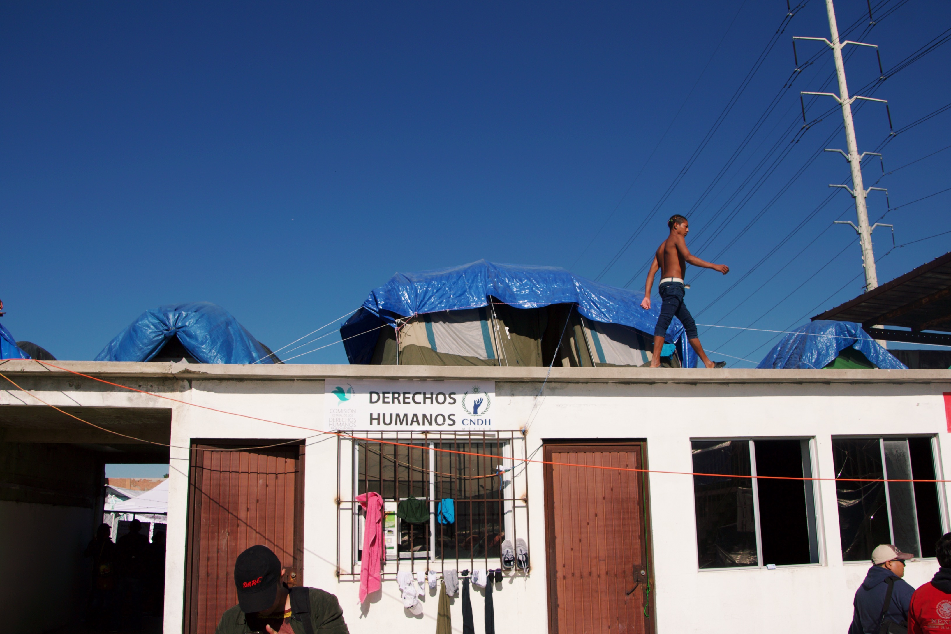 A migrant from Honduras walks by his family's tent on a roof above a portion of El Barretal, the main shelter for migrants who arrived in two caravans from Central America into Tijuana, Mexico in November. The sign below him reads 