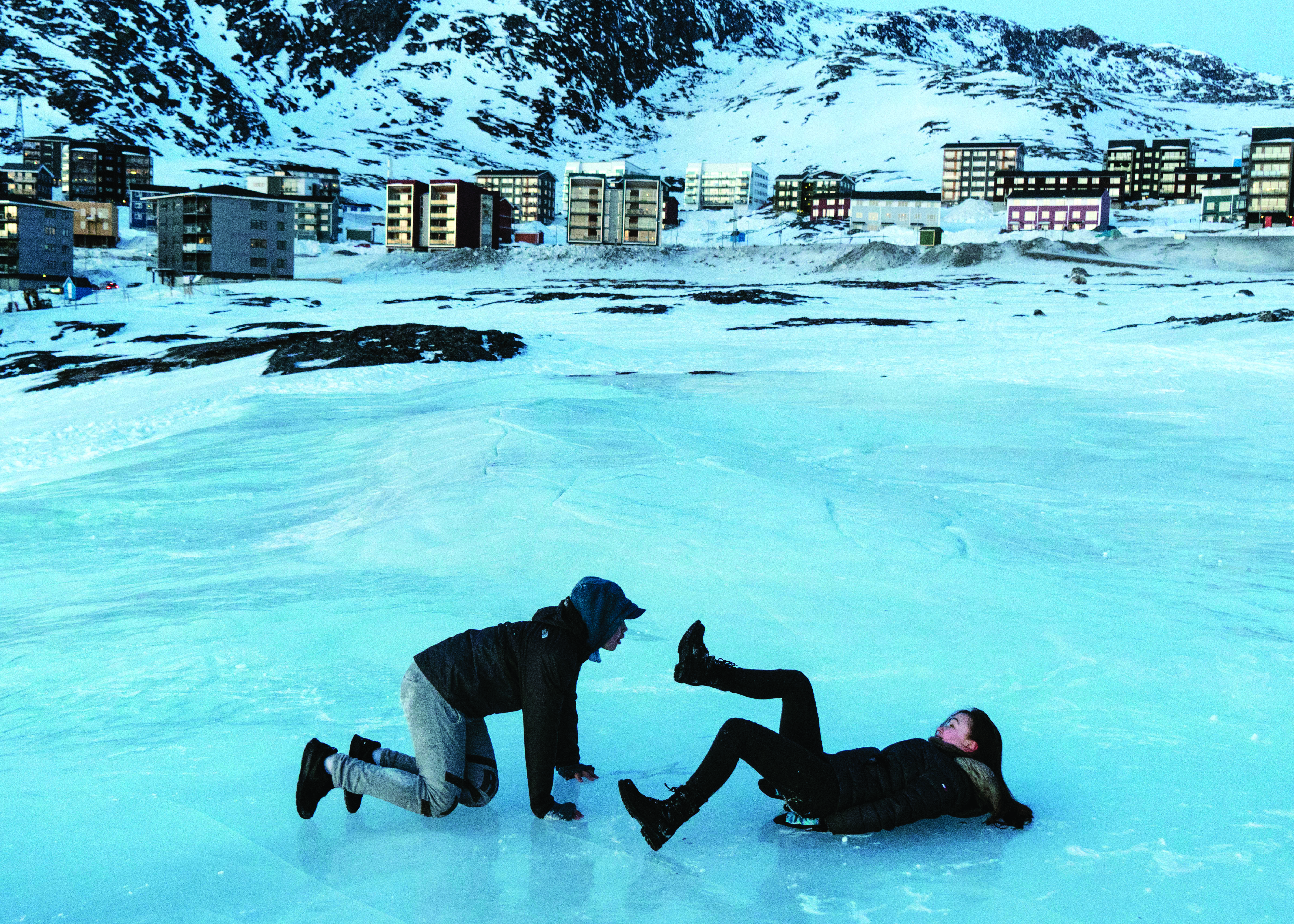 Photo showing two young adults playing on an icy stretch in front of a Greenlandic village (hi res version)