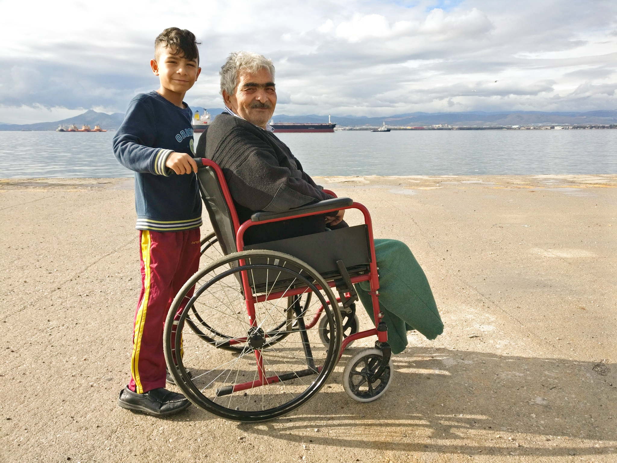 A grandson pushes his grandfather along the water.